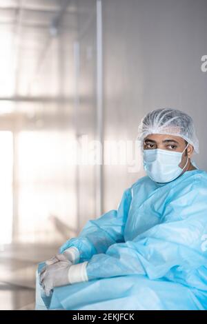 Exhausted surgeon in protective workwear leaning against wall in corridor in front of camera and having little rest before next operation Stock Photo