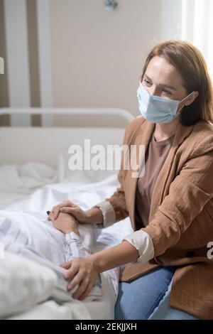 Young emphatic woman in protective mask supporting sick friend with covid during conversation while sitting by her bed in chamber Stock Photo