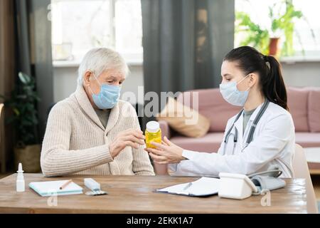 Young female doctor in whitecoat and protective mask holding bottle of pills over table, showing and prescribing them to sick senior man Stock Photo