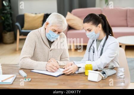 Sick senior man in mask and casualwear signing medical document while sitting by table in front of young female doctor in living-room Stock Photo