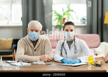 Sick senior male patient and young brunette female doctor in protective masks sitting by table next to one another and looking at you Stock Photo