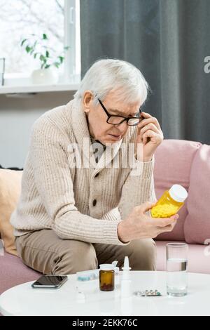 Senior man in casualwear and eyeglasses sitting on couch in living-room, holding bottle of pills over table and reading contains of tablets Stock Photo
