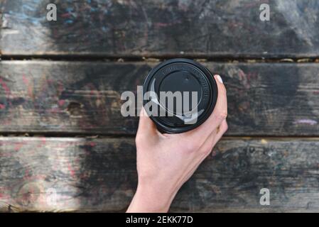 top view of the hands of an unrecognizable caucasian woman taking a disposable paper cup of coffee. On the top of an aged wooden table in a green park Stock Photo