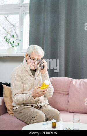 Senior man with mobile phone sitting on couch, looking at bottle of pills in his hands and consulting with his doctor about how to take them Stock Photo