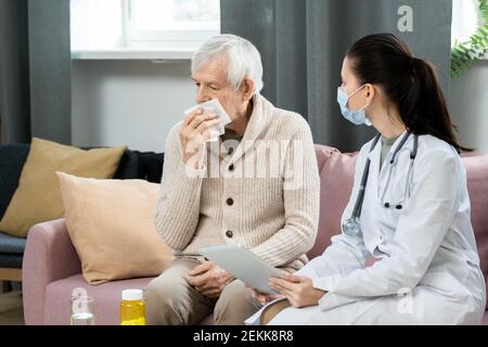 Young female general practitioner with digital tablet looking at sick senior man with handkerchief by his face sitting on couch in front of her Stock Photo