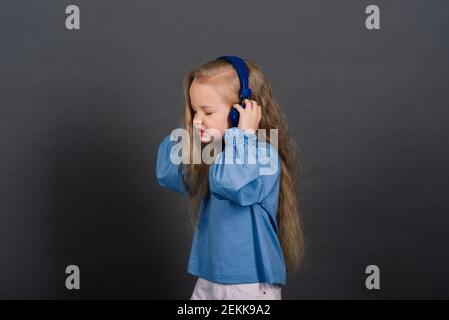 Smiling little girl in headphones on a grey background. The child listens to music, plays and learns online. Electronic digital training. Stock Photo