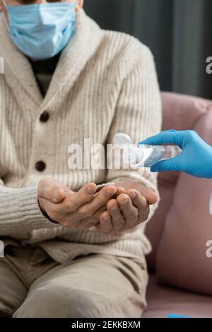 Gloved hand of contemporary general practitioner dropping sanitizer on those of sick senior man sitting on couch in home environment Stock Photo