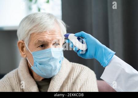 Sick elderly man in protective mask sitting in front of doctor in gloves using contactless thermometer for measuring his body temperature Stock Photo