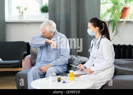 Young female general practitioner with touchpad looking at sick senior man going to cough or sneeze while sitting on couch in front of her Stock Photo