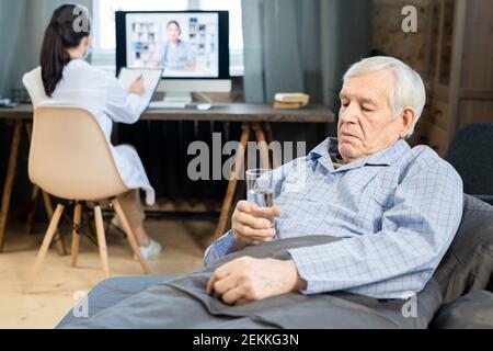 Sick senior male patient in blue pajamas sitting on couch under blanket and looking at glass of water in his hand against doctor working Stock Photo