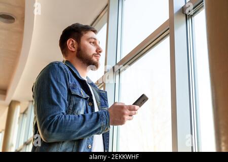 Man use smartphone while standing near window in beautiful modern business center. Young man in denim jacket near panoramic window with phone. Man tex Stock Photo