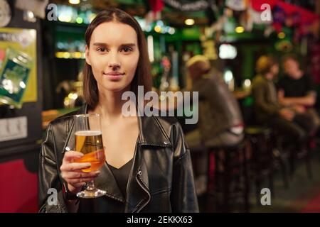 Portrait of young woman holding glass of beer and looking at camera standing in the pub Stock Photo