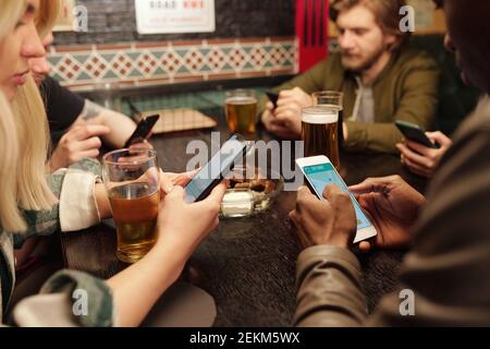 Close-up of friends sitting at the table with glasses of beer and using their mobile phones during meeting in the pub Stock Photo