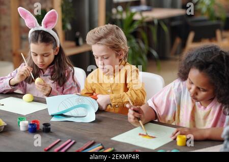 Group of children sitting at the table painting pictures and painting eggs at the table during art lesson Stock Photo