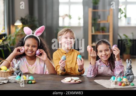 Portrait of happy children holding Easter eggs in their hands and smiling at camera sitting at the table Stock Photo