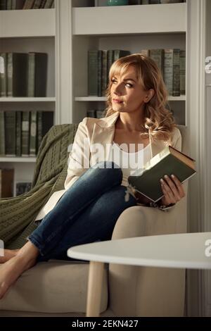 Young woman chilling at home. Girl relaxes and reads a book in the rays of the cozy warm sun. Sitting on retro chair near the window in light her livi Stock Photo