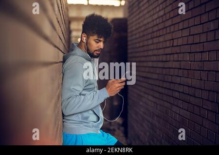 Young serious bearded afro american man with earphones leaning against the wall listening music and holding a mobile between two street walls outside. Stock Photo