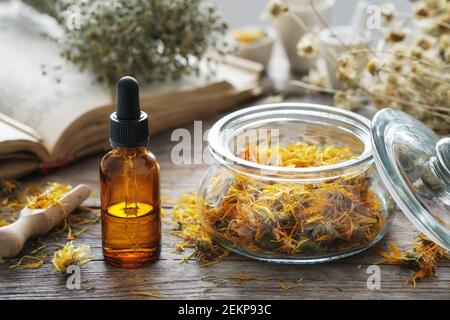 Dropper bottle of calendula infusion or oil, jar of dried marigold flowers, old recipes book and chamomile bunch on background. Alternative medicine. Stock Photo