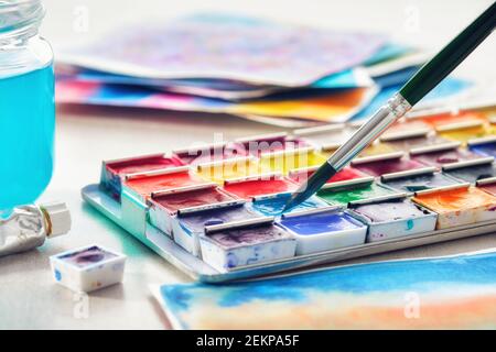 Set of watercolor paints, paintbrush, glass of water and paper sheets of watercolor paintings on background. Selective focus. Stock Photo
