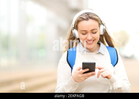 Happy student wearing headphones listening to music on smart phone walking in the street Stock Photo