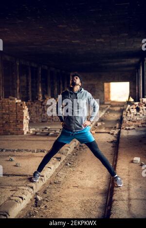 Portrait of active motivated afro-american young attractive athletic man with earphones standing and doing stretching workout with legs open inside of Stock Photo