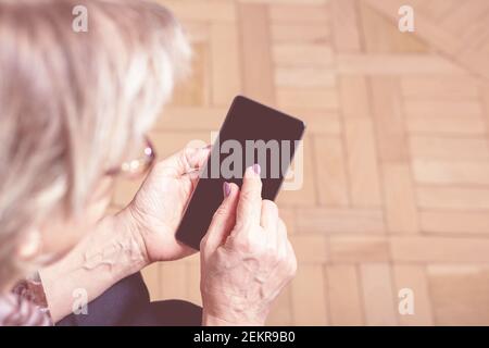 Elderly womans hand holding mobile cell phone. Over shoulder. Old woman looking at cell phone. Close-up. Blanck mobile phone screen. Display media.  Stock Photo