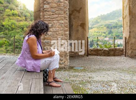 A brunette woman writes postcards while sitting on the steps of an ancient castle in Dolceacqua, Italy. Stock Photo