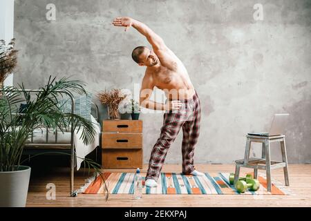 The young man goes in for sports at home. Sportsman with black hair in home pajamas  does morning exercises, bends and pulls his hand on carpet in bed Stock Photo