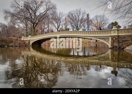 The bow bridge in central park, New York city daylight view in winter with reflection in water , clouds, trees and Manhattan skyline in background Stock Photo