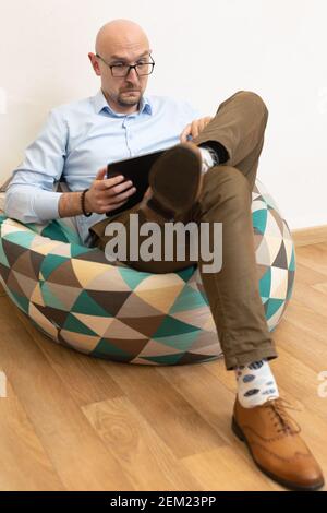 surprised bald businessman in eyeglasses holding digital tablet while sitting on bag chair with dazed look Stock Photo