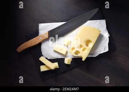 Fresh Maasdam cheese with holes and knife on dark stone plate on wooden background Stock Photo