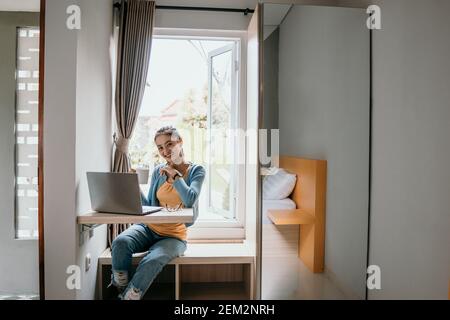 Confident young woman in smart casual wear working on laptop while sitting near window at home