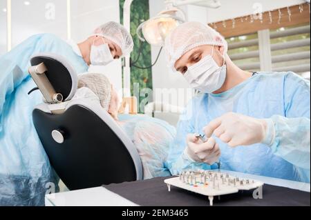 Dentist on foreground, wearing disposable sterile clothes, putting surgical nozzle on drill before implant installation. Patient and assistant on blurred background. Concept of implant placement. Stock Photo