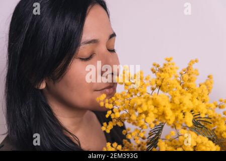 womans day mimosa. closeup of young woman smelling yellow mimosa flowers bouquet on white background smiling with casual clothes Stock Photo