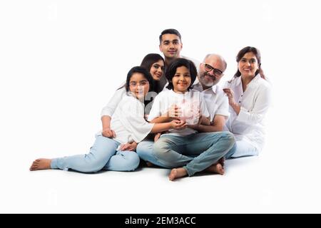 Multigenerational Indian family of six holding piggy bank while wearing white cloths and standing against white wall Stock Photo