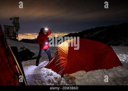Old Climber with glowing headlamp near orange tent in the winter mountain ski resort Shymbulak at dark night sky in Almaty, Kazakhstan Stock Photo