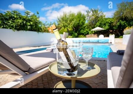 Focus on white wine bottle in a glass container to keep it cool besides some loungers. Outdoors poolside shot out of focus. Stock Photo