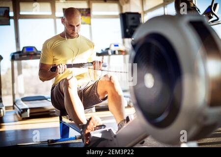 Young handsome muscular active focused man with earphones doing back exercises on the machine while sitting and pulling the bar in the gym. Stock Photo