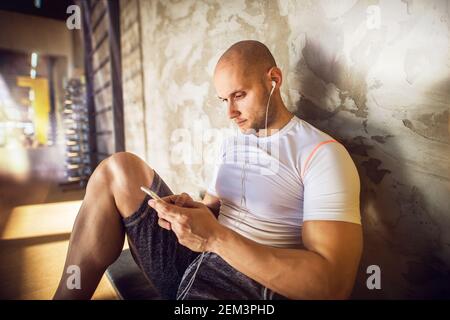 Young muscular shape strong bald man relaxing and resting with music and mobile while sitting leaning against the wall in the gym. Stock Photo