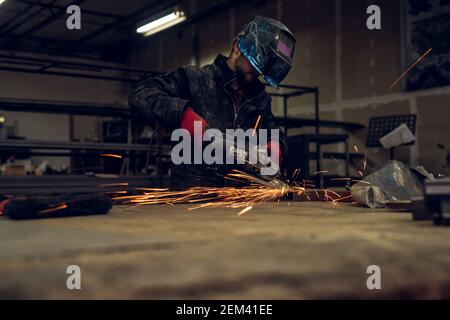 Focused bearded handsome professional industrial worker with protection mask little raised working with an electric grinder in a fabric workshop. Stock Photo