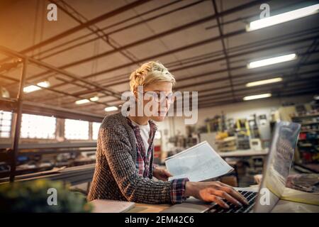 Hardworking professional engineer short hair woman working on a laptop while holding blueprint in another hand in the sunny workshop. Stock Photo