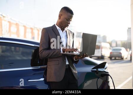 Handsome African American business man in formal suit working with laptop, while standing near charging electric vehicle at outdoor charging station Stock Photo