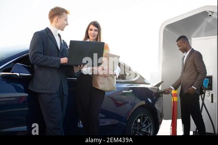 Pleasant 30 aged business people man and woman, using laptop outdoors at urban EV charging station, while their afro american colleague charging Stock Photo