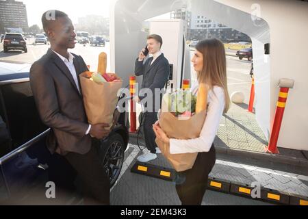 Smiling multiethnic friends at the charging station for electric cars. Caucasian man is charging a car and talking phone, woman and black man with Stock Photo