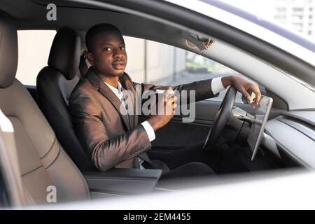 Handsome young dark skinned man in formal wear sitting in his modern electric car and is ready to start charging at EV charging station, while Stock Photo