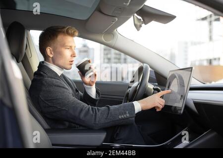 Testing a new electric futuristic car with self driving system. Side view of handsome Caucasian businessman sitting in modern car, drinking coffee to Stock Photo