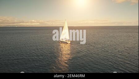 Aerial of yacht cruising in ocean waters at sun with clouds. White sail boat at open sea. Summer vacation at sailboat. Serene seascape with water transport. Cinematic relax and calm waterscape Stock Photo