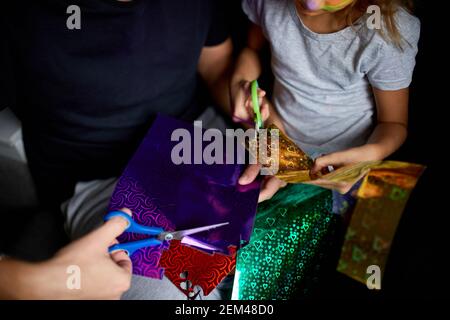 Daughter and father having fun making craft together at home on the sofa, cutting a paper with scissors, dark light, Fathers day, family. Stock Photo