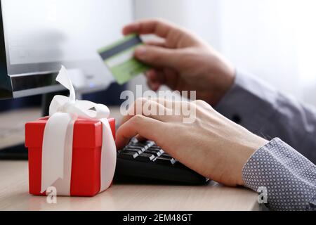 Man with credit card sitting on PC keyboard background and red gift box. Concept of buying a gift, online shopping and payment Stock Photo