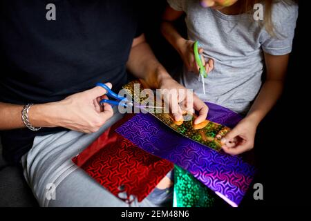 Daughter and father having fun making craft together at home on the sofa, cutting a paper with scissors, dark light, Fathers day, family. Stock Photo